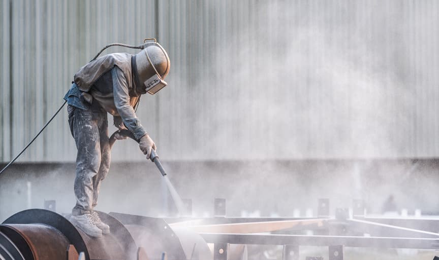 An operator in protective gear blasting a steel fabrication with abrasive media during surface preparation